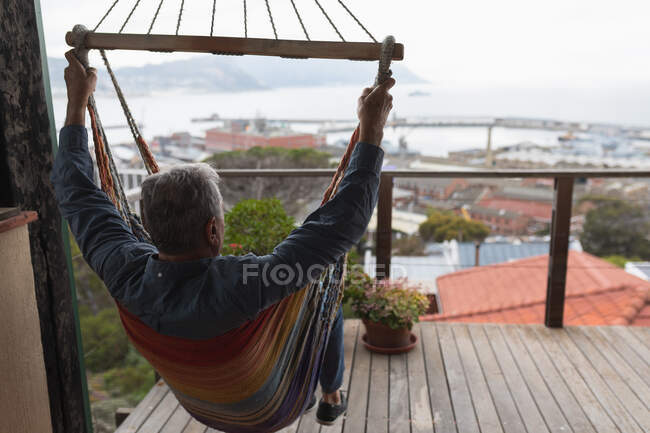 Вид сзади на старшего кавказца, расслабляющегося дома, сидящего в гамаке на балконе, любующегося видом — стоковое фото