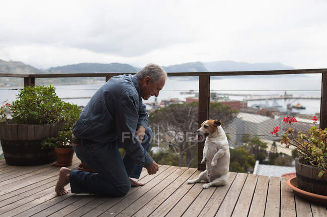 Seitenansicht eines älteren kaukasischen Mannes, der es sich zu Hause gemütlich macht, auf dem Balkon hockt, mit seinem Hund spielt und lächelt — Stockfoto