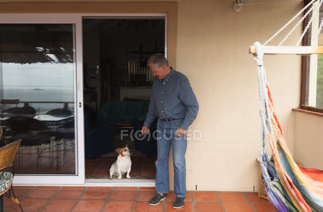 Seitenansicht eines älteren kaukasischen Mannes, der es sich zu Hause gemütlich macht, in einer offenen Tür steht und mit seinem Hund spricht — Stockfoto