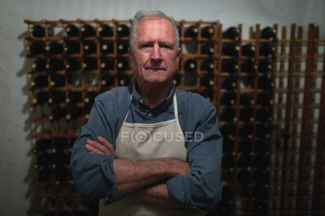Portrait of a senior Caucasian man relaxing at home, standing in his wine cellar, wearing an apron, with arms crossed, looking at camera — Stock Photo