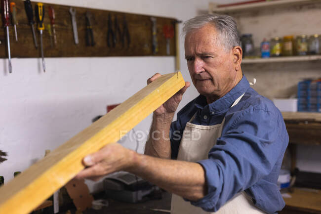 Vue latérale d'un homme caucasien âgé se relaxant à la maison, debout dans son atelier, tenant et vérifiant un morceau de bois, avec des outils accrochés au mur en arrière-plan — Photo de stock