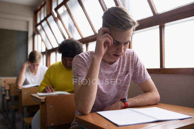 Vorderansicht eines jugendlichen kaukasischen Jungen in einem Klassenzimmer, der konzentriert am Schreibtisch sitzt, während im Hintergrund männliche und weibliche Mitschüler am Schreibtisch sitzen und arbeiten. — Stockfoto
