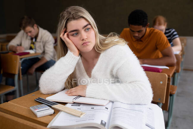 Vue de face d'une adolescente caucasienne dans une classe d'école assise au bureau, la tête penchée sur sa main, regardant ailleurs, avec des camarades de classe adolescents hommes et femmes assis à des bureaux travaillant en arrière-plan — Photo de stock