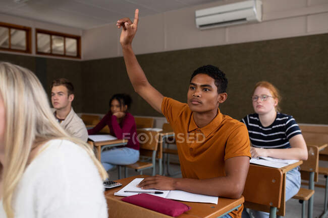 Vue de face d'un adolescent métis dans une classe d'école assis au bureau, levant la main pour répondre à une question, avec des camarades de classe adolescents hommes et femmes assis à des bureaux travaillant en arrière-plan — Photo de stock