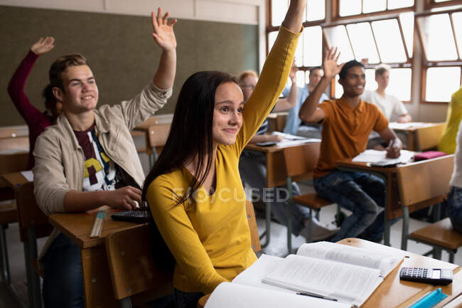 Vista laterale da vicino di un gruppo multietnico di adolescenti sorridenti in una classe scolastica seduti alla scrivania, tutti alzando le mani per rispondere a una domanda — Foto stock