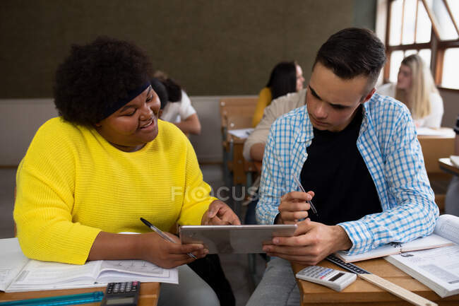 Front view of a teenage Caucasian boy and a teenage African American girl in a high school classroom sitting a desk, using a tablet computer, with teenage classmates sitting at desks working in the background — Stock Photo
