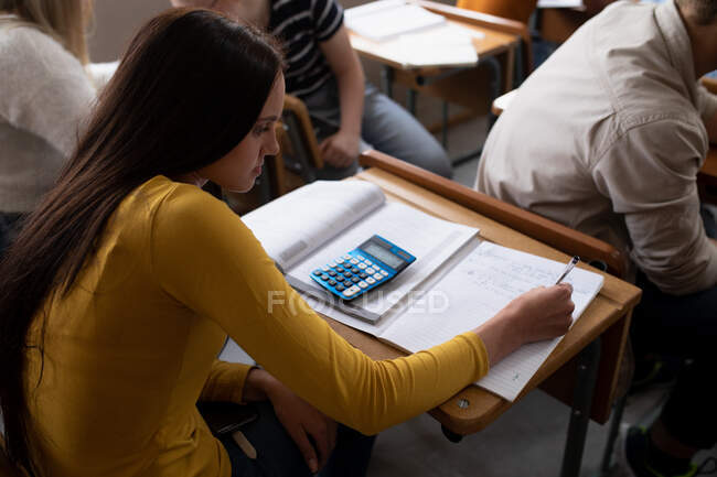 Side view of a teenage Caucasian girl in a school classroom sitting at desk, concentrating and writing, with teenage male and female classmates sitting at desks working in the background — Stock Photo