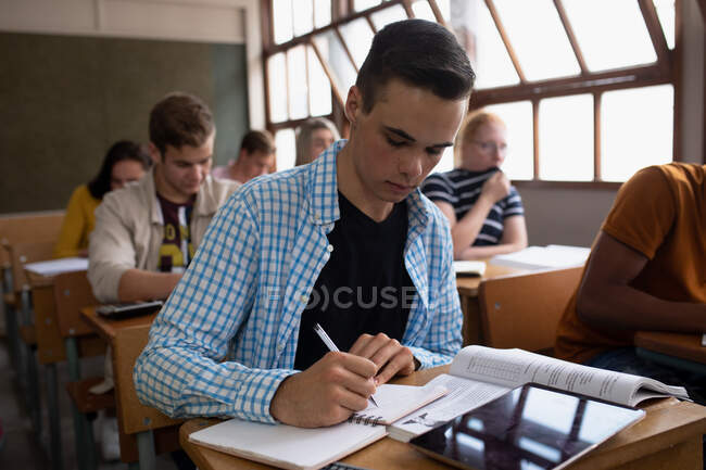 Side view of a teenage Caucasian boy in a school classroom sitting at desk, concentrating and writing, with teenage male and female classmates sitting at desks working in the background — Stock Photo