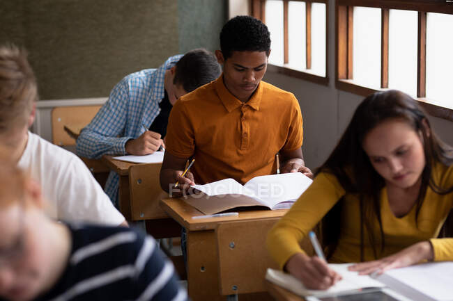 Vista frontal de un adolescente de raza mixta en un aula de la escuela sentado en el escritorio, concentrándose y leyendo, con compañeros de clase adolescentes masculinos y femeninos sentados en escritorios trabajando a su alrededor - foto de stock