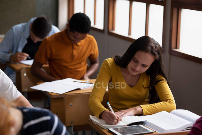 Vue de face d'une adolescente caucasienne dans une classe d'école assise au bureau, se concentrant et écrivant, avec des camarades de classe masculins et féminins adolescents assis à des bureaux travaillant en arrière-plan — Photo de stock