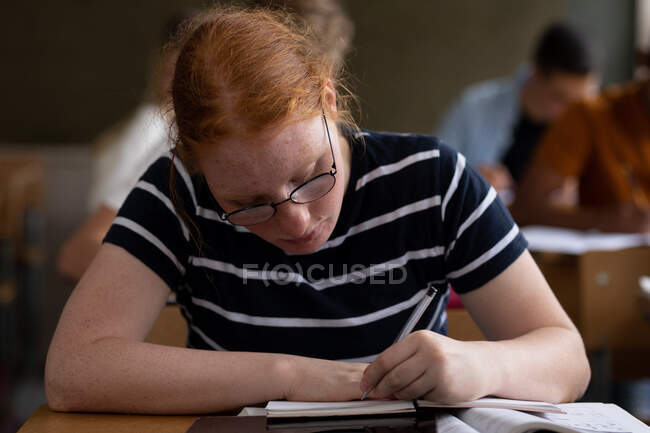 Vista frontal de cerca de una adolescente caucásica en un aula de la escuela sentada en el escritorio, concentrándose y escribiendo, con compañeros de clase adolescentes masculinos y femeninos sentados en escritorios trabajando en el fondo - foto de stock