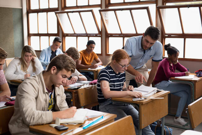 Front view of a Caucasian male high school teacher, standing and talking with a teenage Caucasian girl in a school classroom sitting at desk, with teenage classmates sitting at desks working in the background — Stock Photo