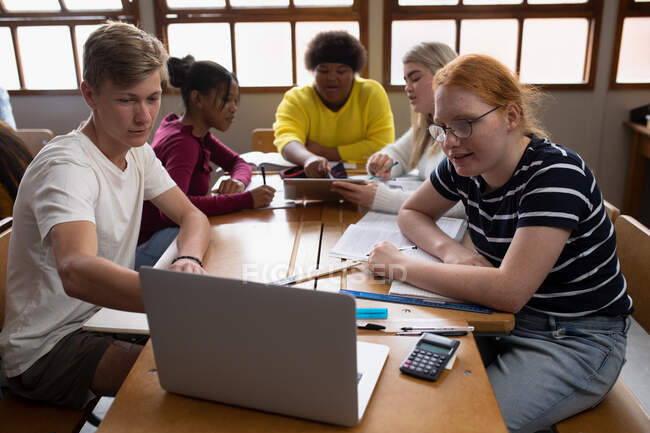 Vista frontal de un grupo multiétnico de adolescentes de secundaria alumnos de escuela masculina y femenina en un aula, sentados en una mesa trabajando juntos, mirando una computadora portátil - foto de stock