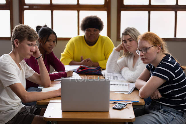 Vue de face d'un groupe multiethnique d'élèves du secondaire, hommes et femmes, dans une salle de classe, assis à une table travaillant ensemble, regardant un ordinateur portable — Photo de stock
