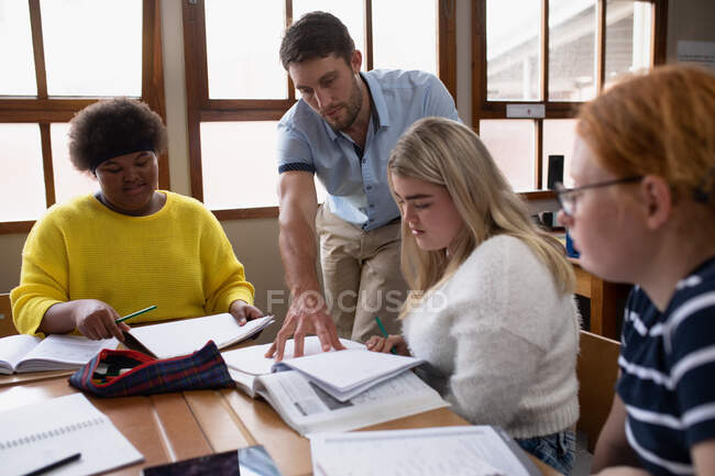 Seitenansicht eines kaukasischen männlichen Gymnasiallehrers, der in einem Klassenzimmer an einem Schreibtisch mit einer multiethnischen Gruppe von Gymnasiastinnen steht und spricht — Stockfoto
