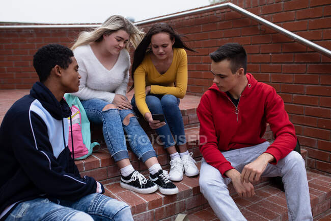 Vista lateral de un grupo multiétnico de adolescentes de secundaria alumnos de escuela masculina y femenina pasando el rato, hablando y mirando un teléfono inteligente juntos, sentados en escalones en sus terrenos escolares - foto de stock
