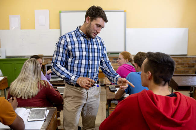 Vista frontal de un maestro caucásico de pie y hablando con un estudiante caucásico en una clase multiétnica de adolescentes en un aula de secundaria sentado en escritorios y trabajando - foto de stock