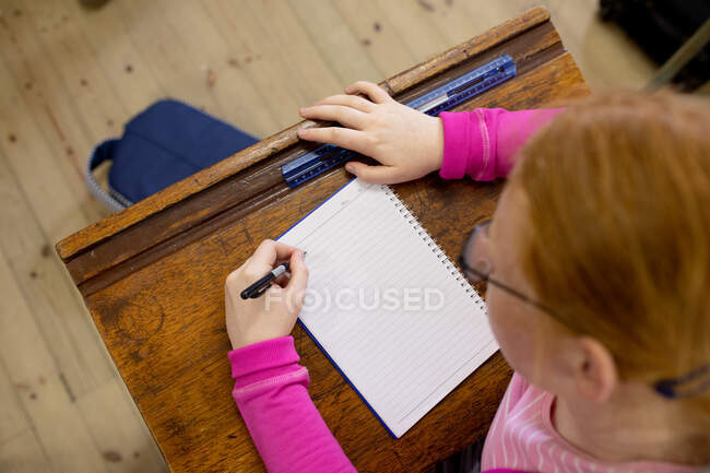 Overhead-Ansicht eines jugendlichen kaukasischen Mädchens in einem Klassenzimmer der High School, das am Schreibtisch sitzt, sich konzentriert und in sein Notizbuch schreibt — Stockfoto