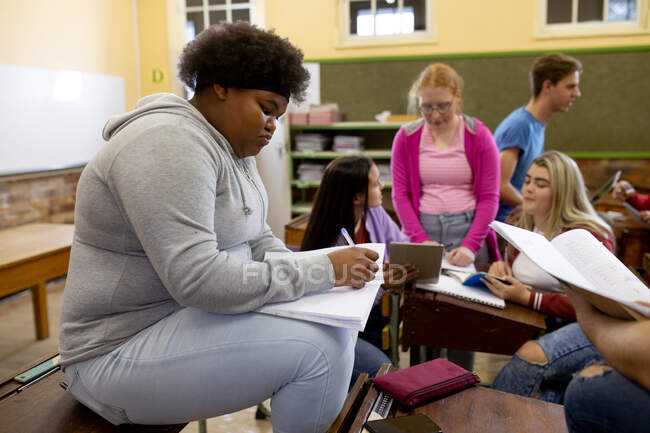 Side view of a teenage African American girl in a high school classroom sitting on a desk, concentrating and writing, with teenage male and female classmates sitting at desks working in the background — Stock Photo