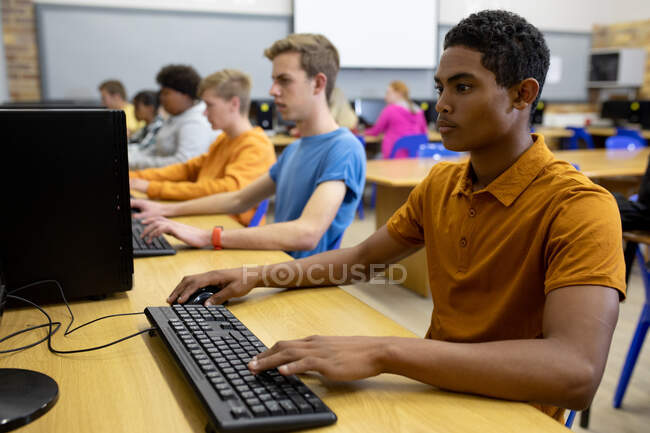 Side view of a mixed race teenage male high school student in a classroom,  working on a computer and concentrating, with other pupils working on  computers in the background — focussed, education -