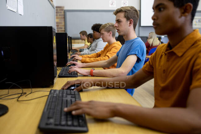 Vista lateral de un grupo multiétnico de adolescentes, hombres y mujeres, estudiantes de secundaria en un aula, trabajando en computadoras y concentrándose - foto de stock
