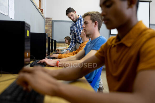 Vue latérale d'un enseignant caucasien homme debout et parlant avec un garçon caucasien adolescent travaillant sur un ordinateur et se concentrant dans une salle de classe, avec des camarades de classe masculins adolescents assis à des bureaux travaillant au premier plan et en arrière-plan — Photo de stock