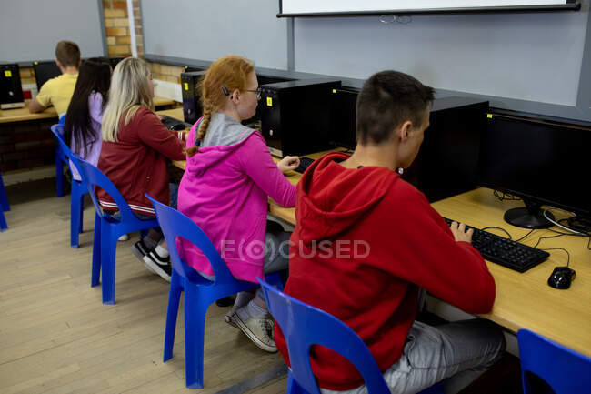 Vista lateral de un grupo multiétnico de adolescentes, hombres y mujeres, estudiantes de secundaria en un aula, trabajando en computadoras y concentrándose - foto de stock