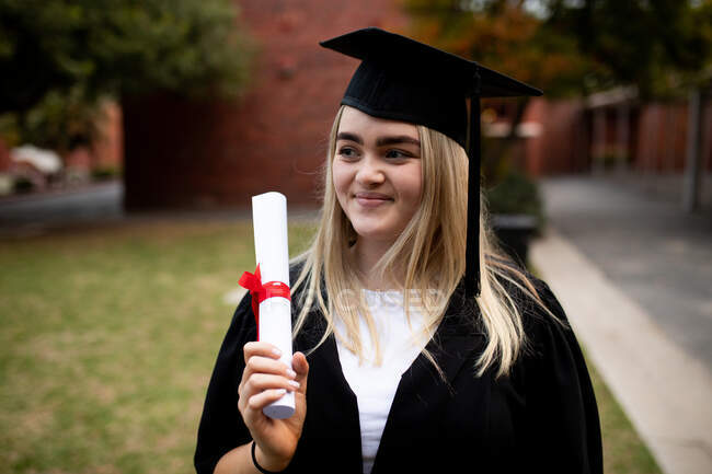Vorderansicht einer kaukasischen Gymnasiastin mit langen blonden Haaren, die eine Mütze und ein Kleid trägt, ein Diplom in der Hand hält und am Tag ihres Abschlusses lächelt — Stockfoto