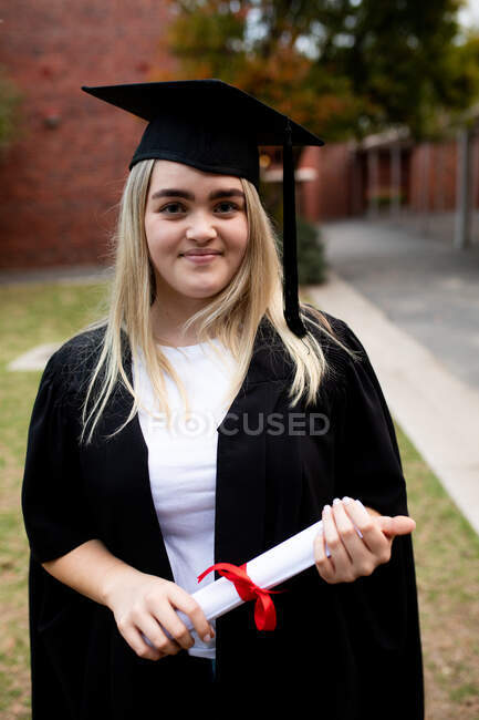 Ritratto di studentessa caucasica adolescente con lunghi capelli biondi che indossa un cappello e un abito, in possesso di un diploma e guardando la fotocamera e sorridendo il giorno del diploma — Foto stock