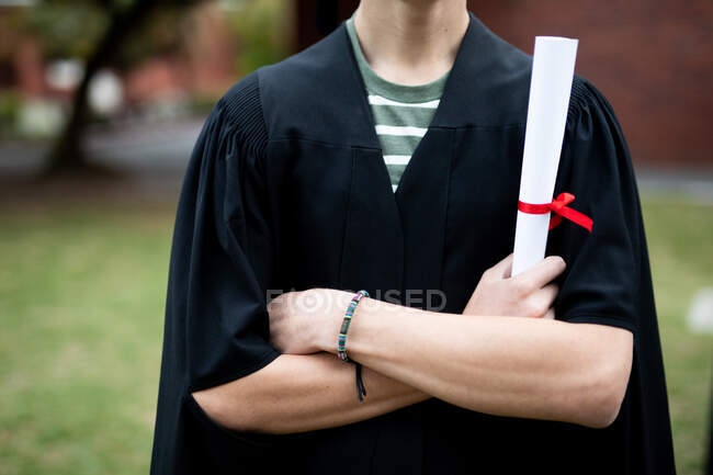 Front view mid section of male high school student wearing a gown, with arms crossed, holding a diploma on his graduation day, with arms crossed — Stock Photo