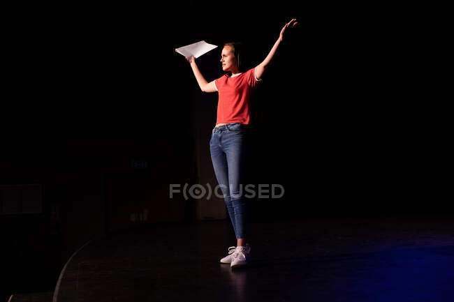 Vista laterale di un'adolescente caucasica del liceo in un teatro vuoto del liceo che si prepara prima di uno spettacolo, in piedi sul palco con le braccia in aria, con un copione in mano e praticando la sua parte — Foto stock