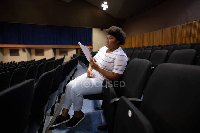 Vista lateral de una adolescente afroamericana en un teatro de secundaria vacío, sentada en el auditorio preparándose para una actuación, sosteniendo un guion y líneas de aprendizaje - foto de stock