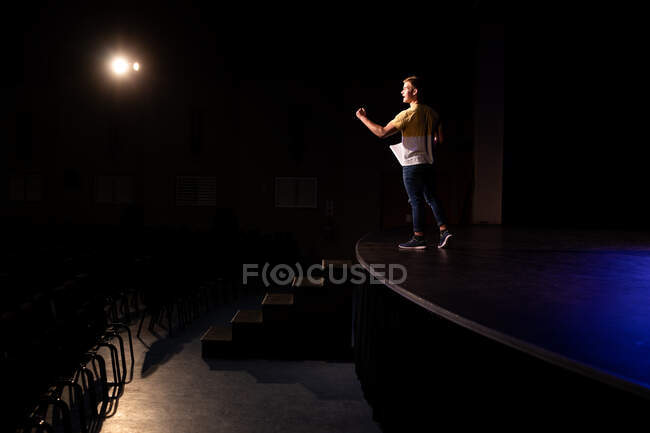 Vista lateral de un adolescente caucásico estudiante de secundaria en un teatro de secundaria vacío preparándose antes de una actuación, de pie en el escenario, sosteniendo un guion y practicando su parte - foto de stock