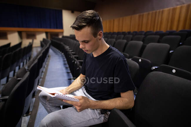 Vista lateral de un adolescente caucásico en un teatro de secundaria vacío, sentado en el auditorio preparándose para una actuación, sosteniendo un guion y líneas de aprendizaje - foto de stock
