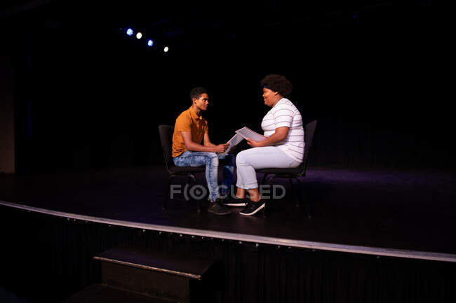 Seitenansicht eines gemischten rassischen Teenagers und eines afroamerikanischen Teenagermädchens, das auf Stühlen in einem leeren Schultheater sitzt und sich vor einer Aufführung vorbereitet, Drehbücher hält und gemeinsam probt — Stockfoto