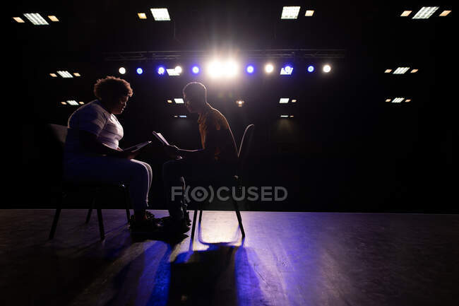 Vista lateral de un adolescente de raza mixta y una adolescente afroamericana estudiante de secundaria sentada en sillas en un teatro escolar vacío preparándose antes de una actuación, sosteniendo guiones y ensayando juntos - foto de stock