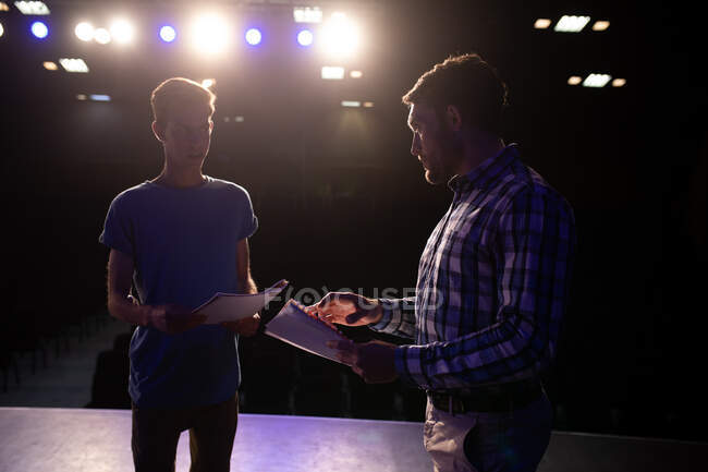 Side view of a Caucasian teenage male high school student and a Caucasian male teacher standing on the stage in an empty school theatre preparing before a performance, holding scripts and practicing together, backlit by spotlights — Stock Photo