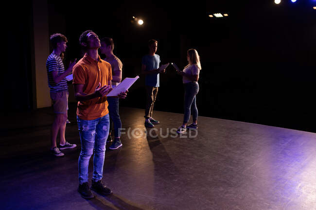 Side view of a mixed race teenage male high school student in an empty school theatre during rehearsals for a performance, standing on the stage holding a script and practicing his part, with other pupils holding scripts in the background — Stock Photo