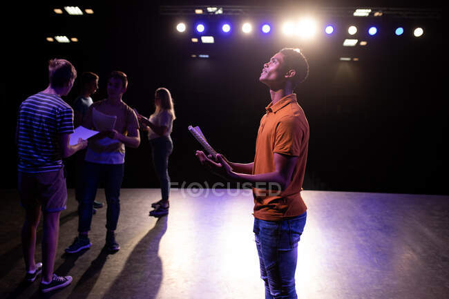 Side view of a mixed race teenage male high school student in an empty school theatre during rehearsals for a performance, standing on the stage holding a script and practicing his part, with other pupils holding scripts in the background — Stock Photo