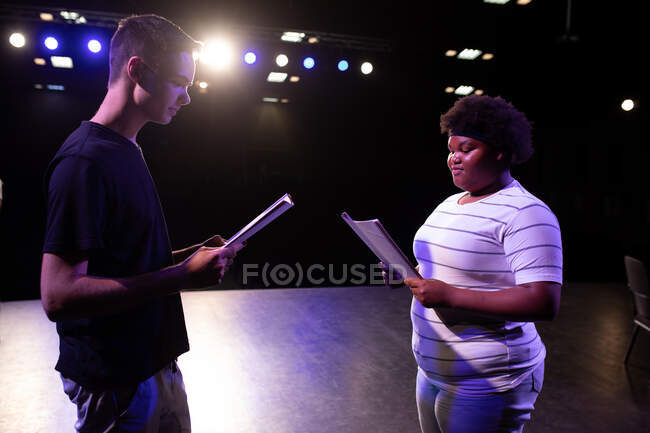Vista lateral de una adolescente afroamericana y un adolescente caucásico estudiante de secundaria en un teatro escolar vacío durante los ensayos para una actuación, de pie en el escenario sosteniendo guiones y practicando sus partes - foto de stock