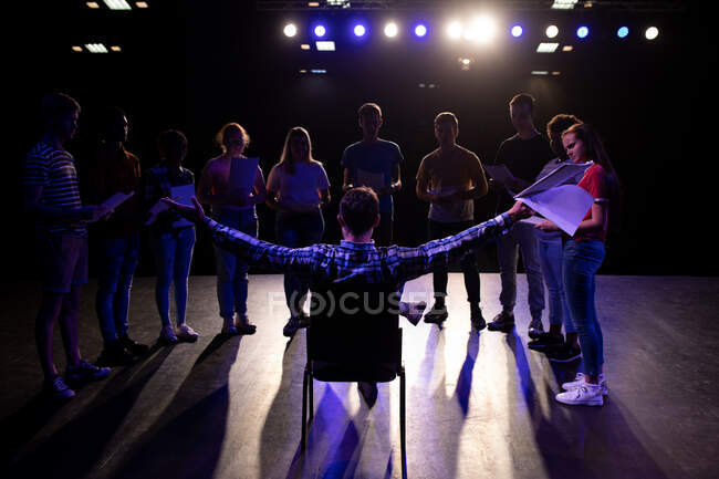Rückansicht eines kaukasischen männlichen Gymnasiallehrers, der auf einem Stuhl auf der Bühne eines leeren Schultheaters während der Proben für eine Aufführung sitzt, ein Drehbuch in der Hand und die Arme ausgestreckt — Stockfoto