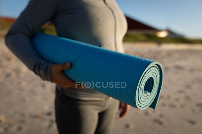 Вид збоку посередині жінки, в сірому костюмі, тримає синій йога килимок під рукою, стоїть на сонячному пляжі . — стокове фото