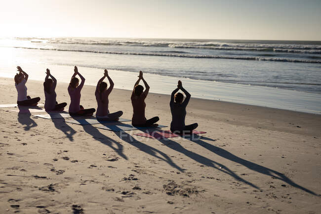 Вид ззаду на багатоетнічну групу друзів, які насолоджуються фізичними вправами на пляжі в сонячний день, практикують йогу, сидячи в положенні йоги, виходять на море, в силуеті . — стокове фото