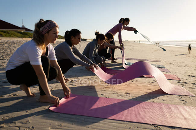 Vista lateral de un grupo multiétnico de amigas disfrutando de la playa en un día soleado, preparando colchonetas de yoga para practicar yoga. - foto de stock