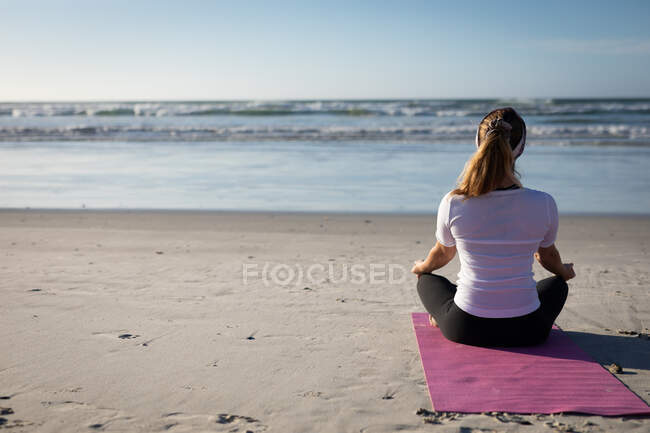 Visão traseira de uma mulher caucasiana que gosta de se exercitar em uma praia em um dia ensolarado, praticando ioga sentado na posição de ioga, meditando na posição de lótus, de frente para o mar. — Fotografia de Stock