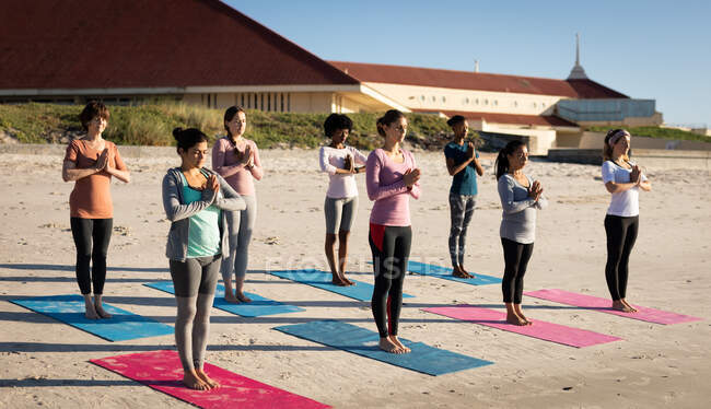 Vista frontal de un grupo multiétnico de amigas disfrutando haciendo ejercicio en una playa en un día soleado, practicando yoga, de pie en posición de yoga. - foto de stock