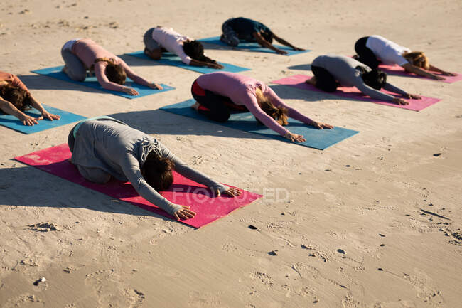 Vista lateral de un grupo multiétnico de amigas disfrutando haciendo ejercicio en una playa en un día soleado, practicando yoga, sentadas en posición de yoga. - foto de stock