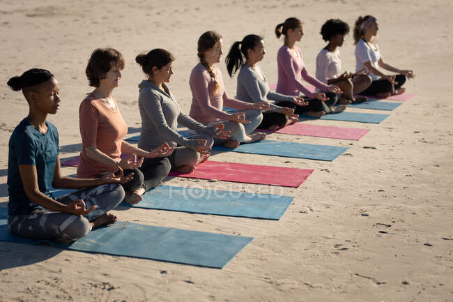 Seitenansicht einer multiethnischen Gruppe von Freundinnen, die an einem sonnigen Tag am Strand Sport treiben, Yoga praktizieren, in Yogaposition mit geschlossenen Augen und Mudra-Händen sitzen, meditieren. — Stockfoto