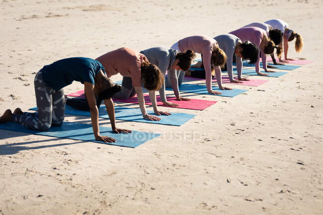 Vista lateral de un grupo multiétnico de amigas disfrutando de hacer ejercicio en una playa en un día soleado, practicar yoga, arrodillarse en posición de yoga. - foto de stock