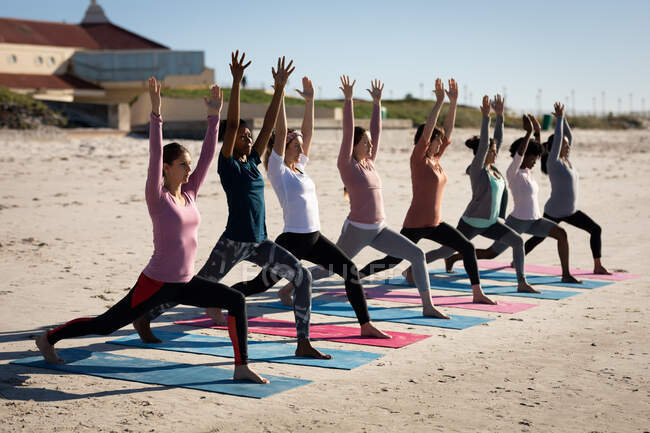 Vista laterale di un gruppo multietnico di amiche che si esercitano su una spiaggia in una giornata di sole, praticano yoga, in piedi in posizione yoga. — Foto stock
